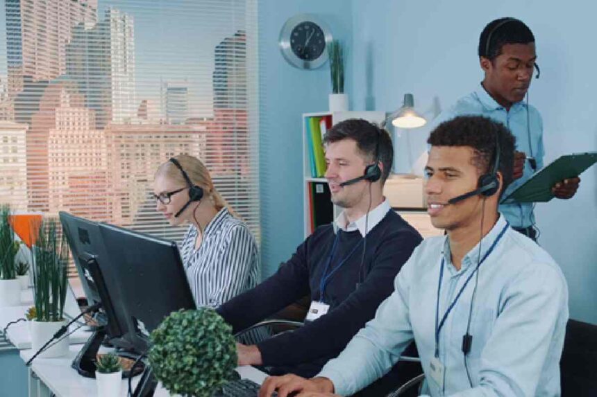 Diverse customer support team wearing headsets and working at computers in a bright office with city views.